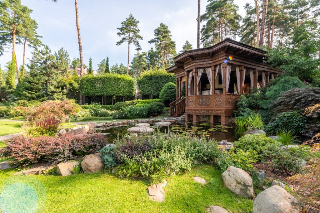 gazebo-near-lake-with-rocks-near-plants-7174103 A peaceful wooden gazebo in a vibrant garden featuring a lush pond and diverse plants in summer.